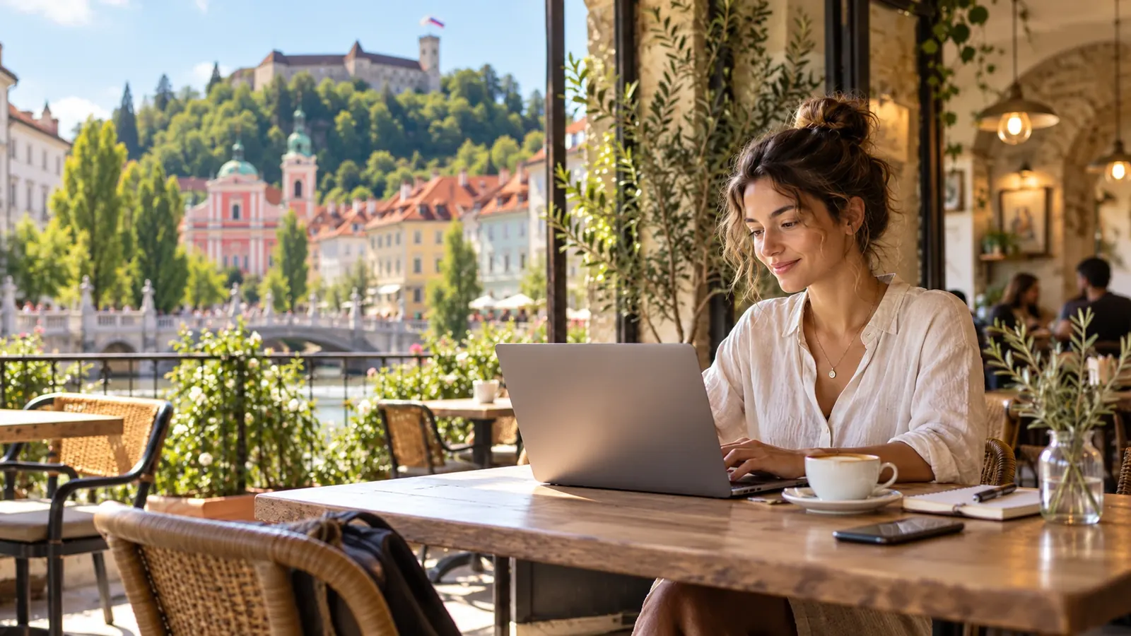 A woman working on a laptop at a riverside café in Ljubljana with a view of Ljubljana Castle